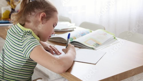 A cute blonde school-age girl is doing her homework sitting at the kitchen table. The child does written assignments in a workbook. The concept of online and home education. photo