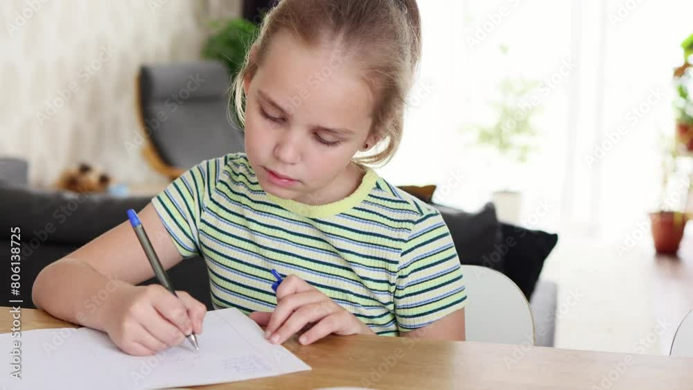 A pretty blonde school-age girl is doing her homework sitting at the kitchen table. She performs written assignments in a workbook. The concept of online and home education. A pretty blonde school-age girl is doing her homework sitting at the kitchen table. She performs written assignments in a workbook. The concept of online and home education.