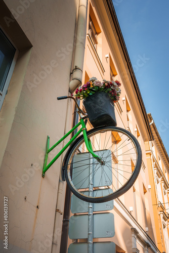Urban installation with a green bicycle on a wall, adorned with colorful flowers above gray electrical boxes. Possibly located in old town Riga, under a clear sky.
