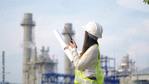 A female engineer in a hard hat and reflective vest holds blueprints and communicates via walkie-talkie at an industrial power plant.

