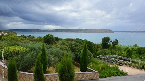 Coastline of a Croatian bay near Razanac town, northern Dalmatia, with lovely tall cypress trees and other foliage, settlements in background, low decorative wall in forefront. Cloudy summer weather. 