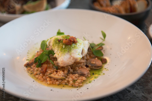 Seared Fish with Herb Sauce on a Bed of Israeli Couscous, Clams, and Garnished with Fresh Herbs, Capturing the Essence of Gourmet Seafood Cuisine