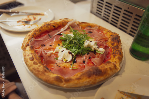Prosciutto and Arugula Pizza Topped with Fresh Burrata Cheese, Balsamic Reduction, and Wood-Fired Crust, Plated on a Rustic White Table with a Side of Water