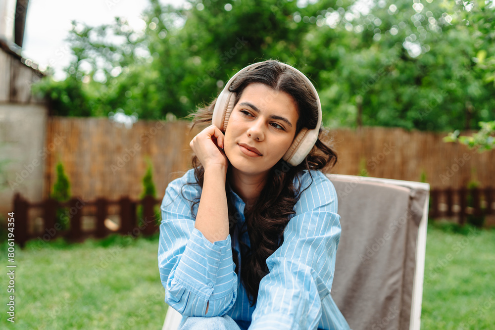 One young caucasian woman is enjoying music on her wireless headphones while laying on easy chair in her backyard One young caucasian woman is enjoying music on her wireless headphones while laying on easy chair in her backyard