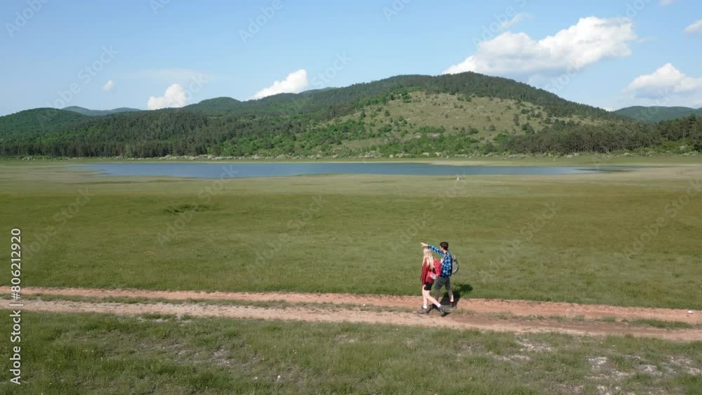 Hiking couple enjoy walking along a trail near blue mountain lake, exploring beautiful nature on a sunny summer day, aerial view. Outdoors recreation concept. Hiking couple enjoy walking along a trail near blue mountain lake, exploring beautiful nature on a sunny summer day, aerial view. Outdoors recreation concept.
