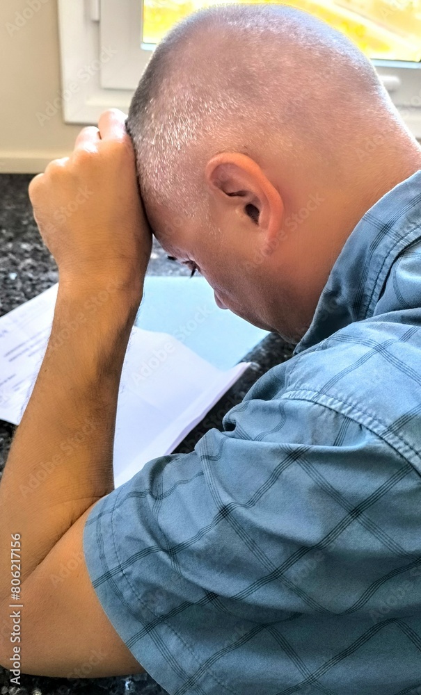 Young adult man, from behind, sitting at the desk, with his hand in a fist on his forehead. Despair. Depression. Economic or financial problems. Not making ends meet financially. Complicated life Young adult man, from behind, sitting at the desk, with his hand in a fist on his forehead. Despair. Depression. Economic or financial problems. Not making ends meet financially. Complicated life