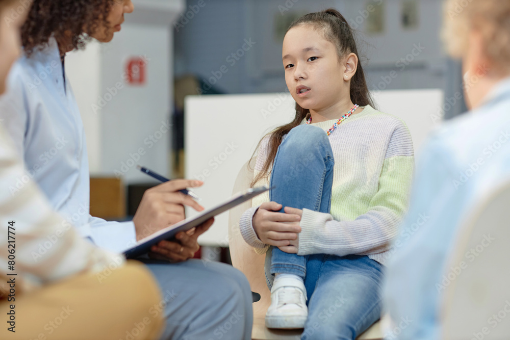 Fototapeta premium Portrait of teen Asian girl sharing story with therapist during support group meeting for childrenwith copy space