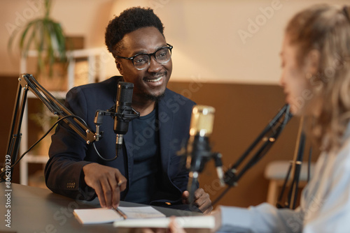 Medium shot of cheerful African American man wearing glasses and black suit talking to unrecognizable female guest sitting at table during podcast in studio