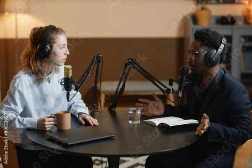 Side view of Caucasian woman with cup talking to gesturing Black man in suit during podcast, both wearing headphones while sitting at round table in studio