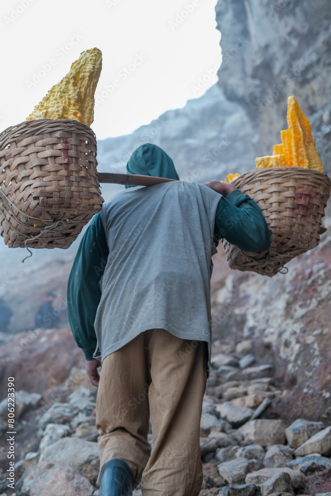 Sulfur miner climbs rocky trail at Kawah Ijen, Banyuwangi, carrying heavy sulfur chunks through extreme volcanic conditions filled with intense sulfur fumes and rugged terrain.