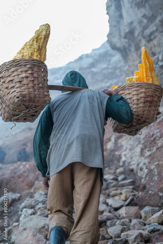Obraz na płótnie Sulfur miner climbs rocky trail at Kawah Ijen, Banyuwangi, carrying heavy sulfur chunks through extreme volcanic conditions filled with intense sulfur fumes and rugged terrain. w salonie