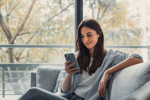 Happy pleasant millennial woman relaxing on comfortable couch, holding smartphone in hands. Smiling young lady chatting in social networks, watching funny videos, using mobile applications at home..