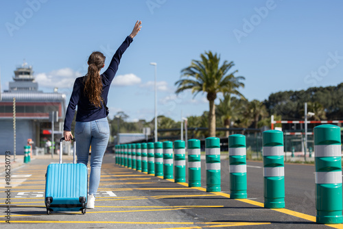 happy girl with her hand raised while walking with her blue carry-on suitcase on the way to the airport to go on summer vacation photo