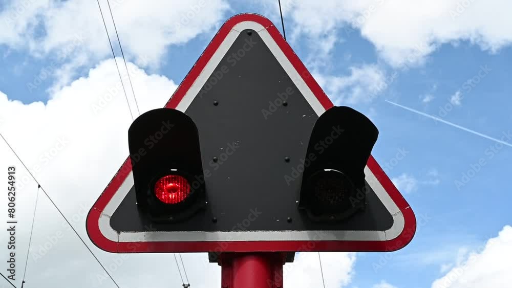 Red railroad crossing lights are positioned within a black triangle that has a red and white frame, set against a blue sky dotted with white clouds and are blinking,