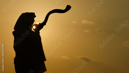 Silhouette of a Jewish man wearing a tallit and tefillin, blowing a long, curly shofar made from the horn of a kudu antelope at sunrise in Israel..