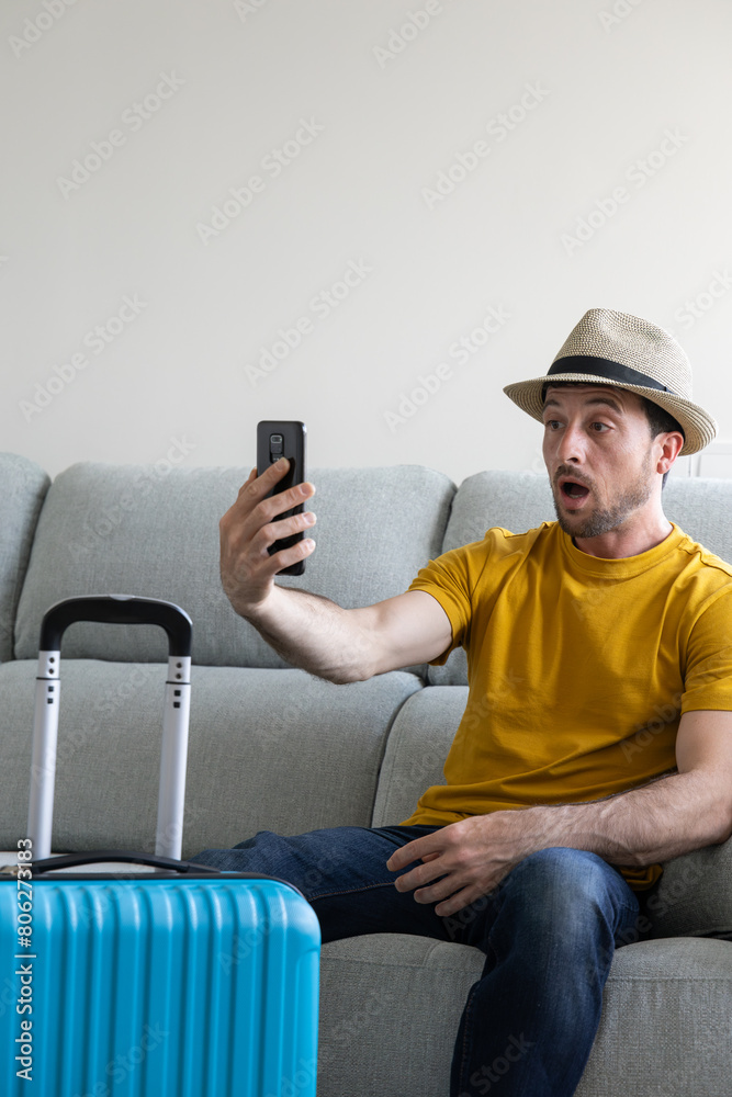 man sitting on the sofa with his blue suitcase ready taking a selfie with a surprised face for not knowing the destination of his summer trip man sitting on the sofa with his blue suitcase ready taking a selfie with a surprised face for not knowing the destination of his summer trip