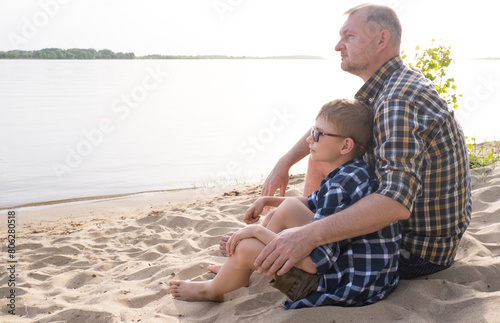 Obraz na płótnie Father's day concept. Father and son sit together on the river bank and look into the distance. w salonie