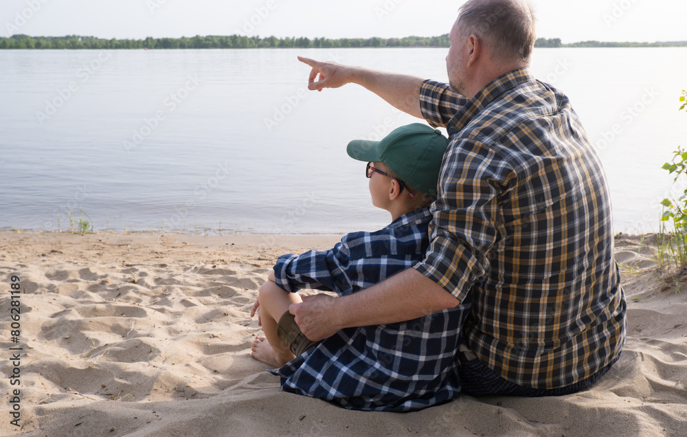 Father's day concept. Rear view of father and son sitting together on the river bank and looking at the water.
