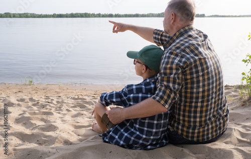 Obraz na płótnie Father's day concept. Rear view of father and son sitting together on the river bank and looking at the water. w salonie