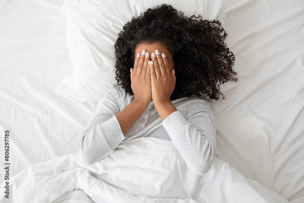 A woman is shown laying in bed, with her face covered by her hands. She appears to be shielding herself, possibly in a state of distress or fatigue. A woman is shown laying in bed, with her face covered by her hands. She appears to be shielding herself, possibly in a state of distress or fatigue.