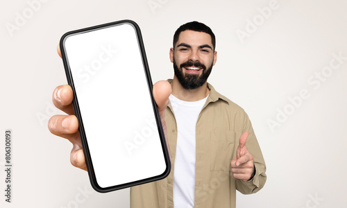 Bearded man is standing, holding a smart phone with white blank screen in his hand. The phone appears to be in use, as he is looking at the screen intently.