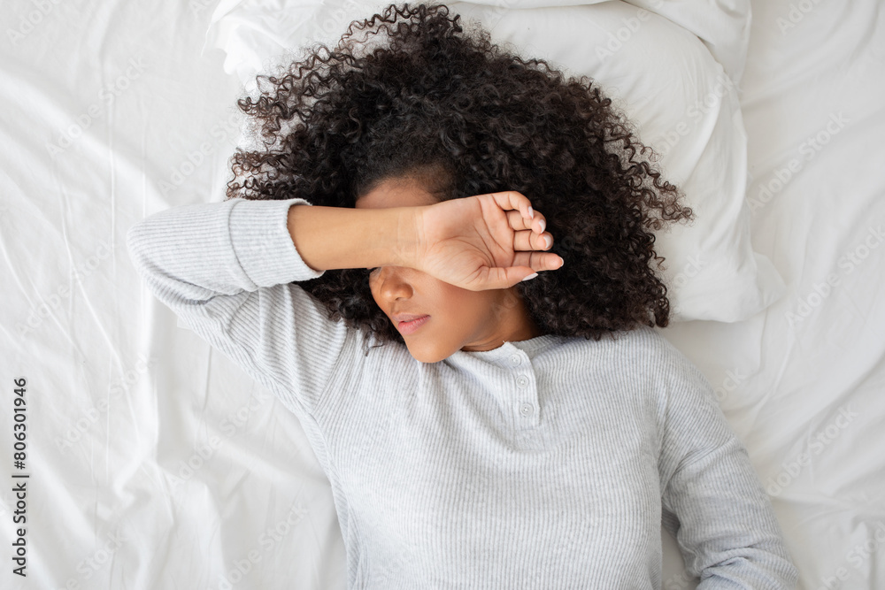 A young woman is lying on a white bedspread, covering her face with her arm as she wakes up. The natural morning light suggests the start of a new day