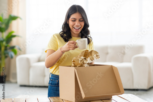 Bild auf Leinwand A cheerful young middle eastern woman is standing in a bright, cozy room, enjoying the process of unpacking parcel, holding cup