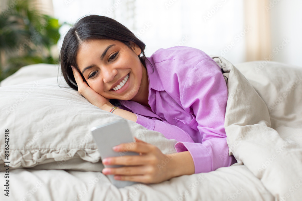 A cheerful young middle eastern woman lies comfortably in bed, wearing a pink shirt, as she enjoys browsing on her smartphone in the softly lit calmness of the morning A cheerful young middle eastern woman lies comfortably in bed, wearing a pink shirt, as she enjoys browsing on her smartphone in the softly lit calmness of the morning