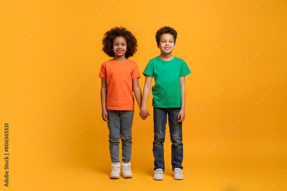 Two African American kids are standing shoulder to shoulder, looking straight ahead. They are both wearing casual clothing and appear relaxed in each others company.