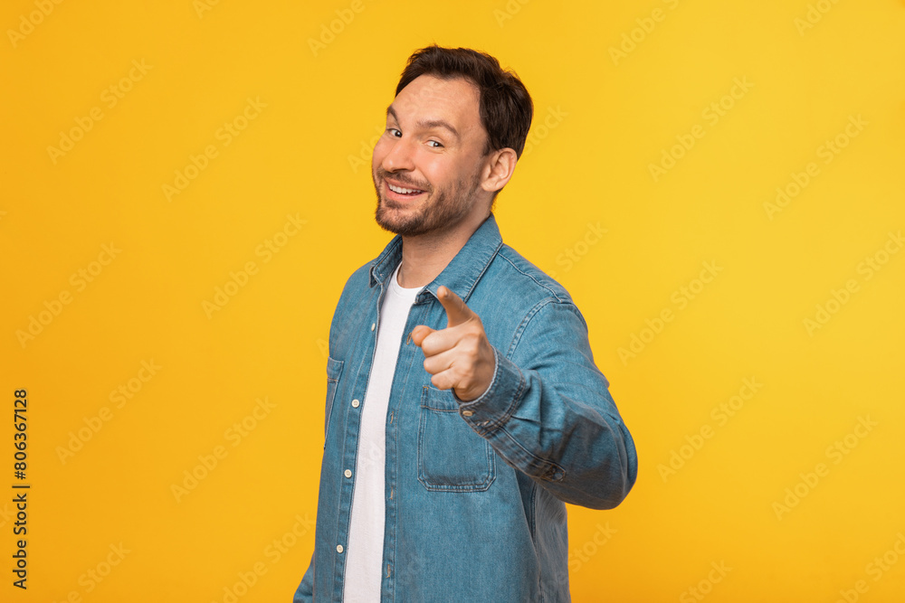 A cheerful man wearing a casual blue denim shirt stands against a vibrant yellow backdrop, pointing directly at the camera with a friendly expression