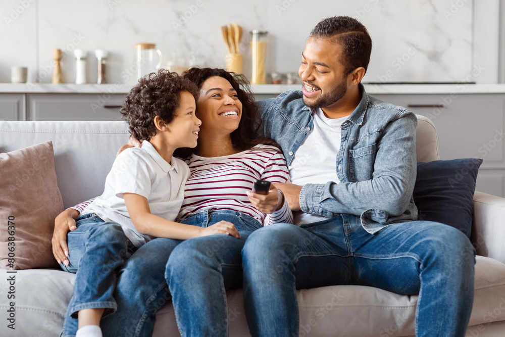 African American man and woman are seated on a couch, with a child between them. The man has short hair, and the woman has long hair. They are all smiling and appear to be engaged in a conversation.