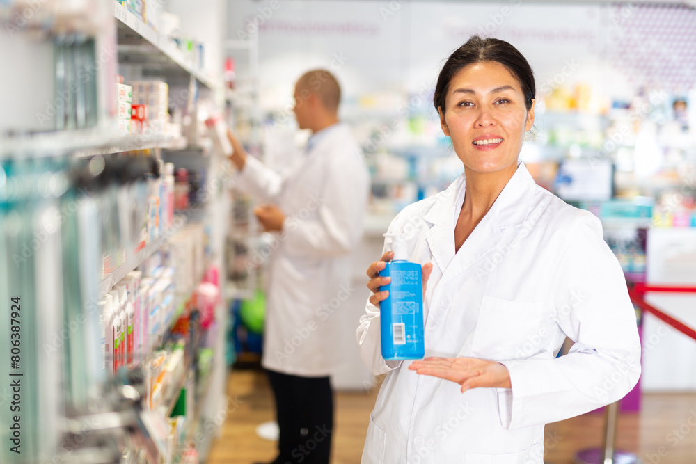 Asian female pharmacist standing in drugstore and holding bottle with sanitizer in hands. Her male colleague standing beside shelf in background.