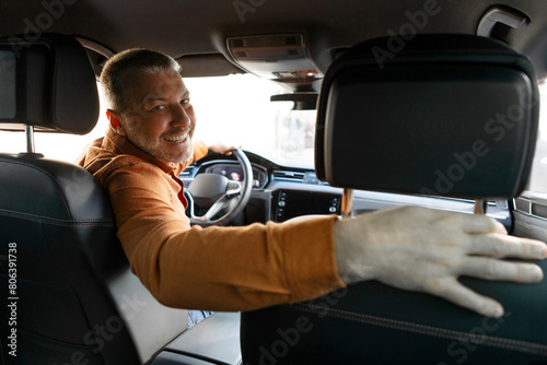 Positive middle aged male driver turning and looking back at camera, sitting in new luxury car, view from rear passenger seat