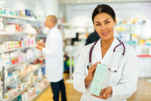 Asian female doctor in white gown standing in drugstore and holding pharmaceutical package in hands. Her male co-worker standing beside shelf in background. photo