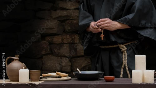 A monk in a black cassock sits at a table in a stone cell and eats soup with bread and onions and drinks wine. photo