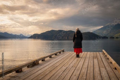 Senior woman gazing out over a lake at sunset from a boat dock. Seen at Harrison Lake, British Columbia, with dramatic clouds and calm waters. photo