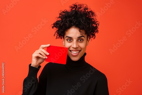 Playful Woman with Curly Hair Holding Red Card, Stylish Casual Attire on Orange Background photo