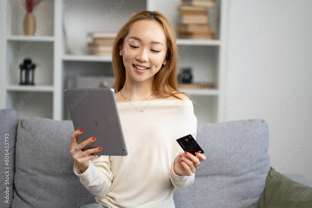 Young Asian Woman Shopping Online On Tablet At Home, Smiling As She Holds A Credit Card, Cozy And Comfortable Living Room Setting Young Asian Woman Shopping Online On Tablet At Home, Smiling As She Holds A Credit Card, Cozy And Comfortable Living Room Setting