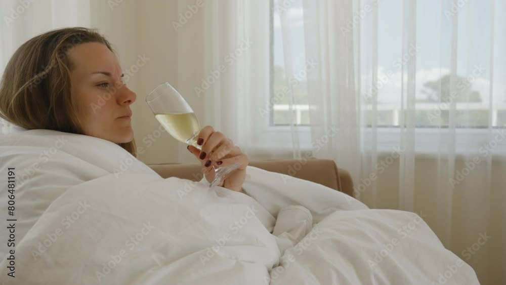 A young woman drinks champagne in a hotel room, wrapped in a white blanket, sitting in an armchair by the window. A young woman drinks champagne in a hotel room, wrapped in a white blanket, sitting in an armchair by the window.