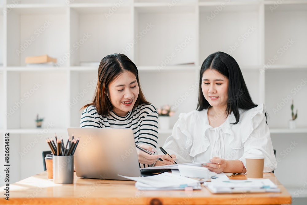 Two women in a contemporary office setting engaged in discussion over work documents with a laptop and stationery on the table. Two women in a contemporary office setting engaged in discussion over work documents with a laptop and stationery on the table.