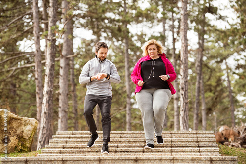 Overweight woman running down the stairs, personal trainer checking her performance. Exercising outdoors for people with obesity, support from friend, fitness coach.