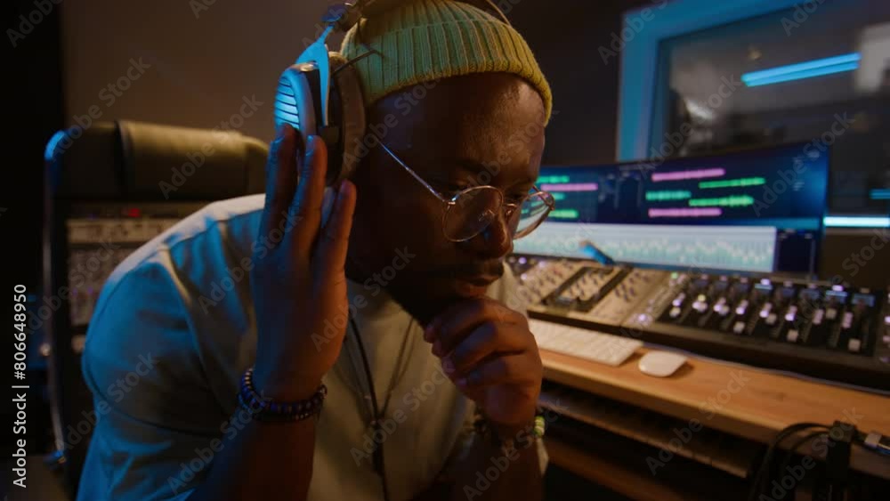 Medium close-up of black male music producer in beanie, glasses, headphones working on new song in studio, listening to track, bobbing head to beat, frowning, concentrated on creative process