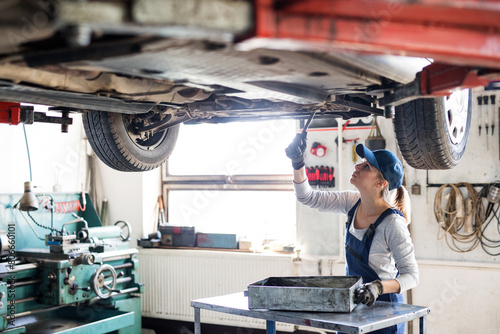 Female auto mechanic elevating car on car lift, changing oil. Beautiful woman working in a garage, wearing blue coveralls.