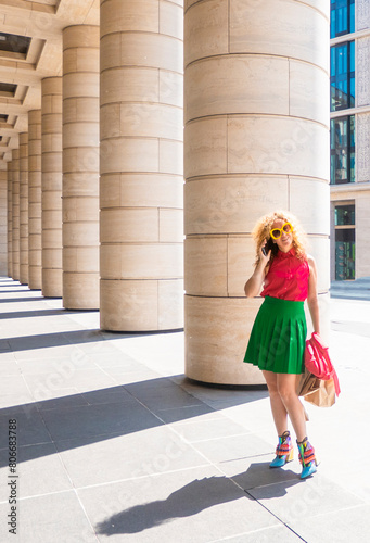 Content fashion female with bag walking on pavement near stone columns in sunlight and talk cell phone. the concept of freedom of tourism and fashion business