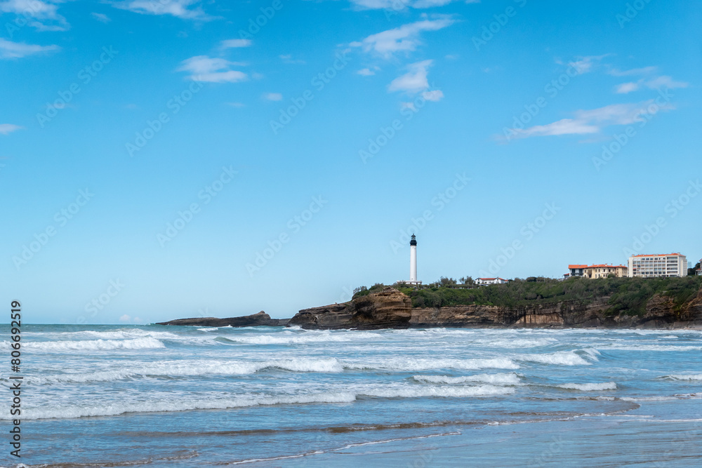 A praia com pequenas ondas, o farol distante em cima de uma grande formação rochosa: A beleza costeira de Biarritz