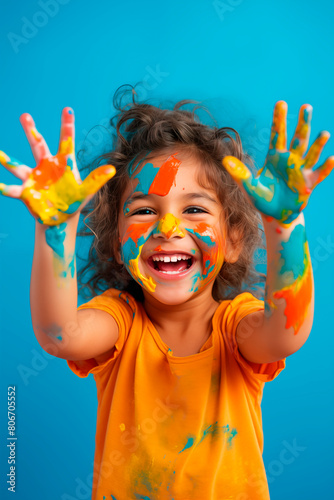 A joyful young girl with hands covered in colorful paint  smiling brightly as she showcases her creative and playful spirit against a vibrant blue background.