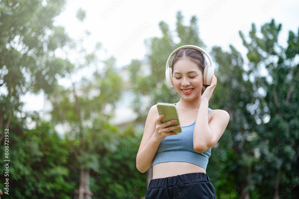 training, fit, fitness, run, runner, smile, player, people, music, relax. A woman is standing in a park, looking at her phone. She is wearing headphones and she is listening to music.