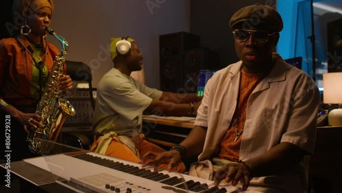 Medium of three African American jazz musicians - black man in cap, sunglasses on keyboard, woman in turban playing saxophone, audio engineer in headphones on mixing board, recording album in studioMe photo