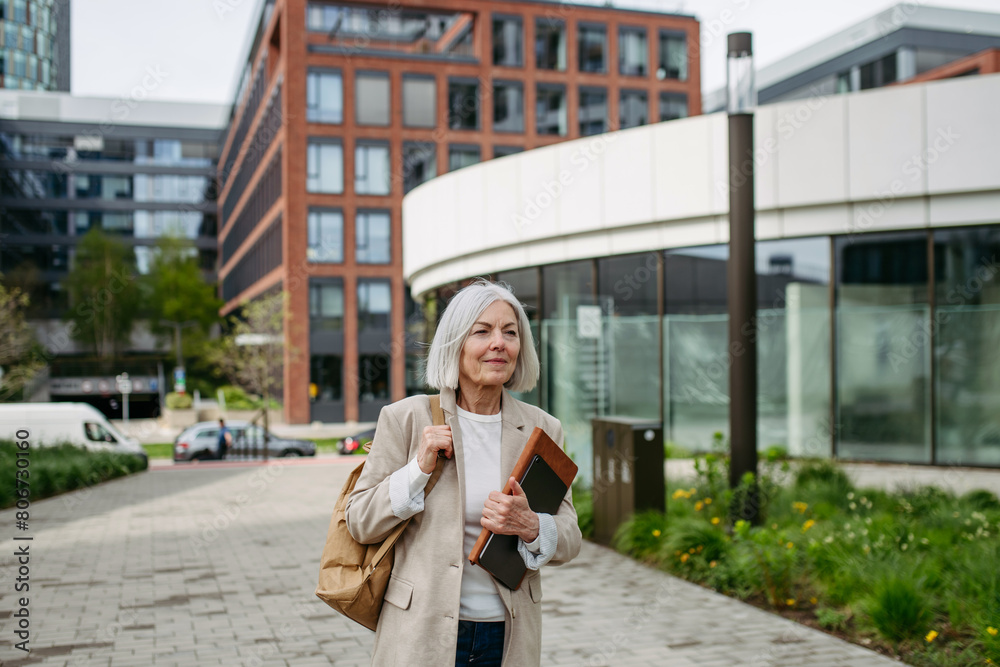 Mature businesswoman going home from work, enjoying beautiful weather and free time. Beautiful older woman with gray hair walking down city street.