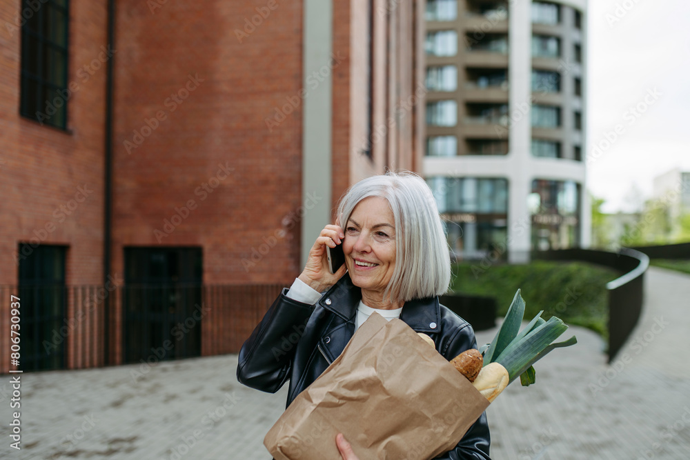 Mature woman making phone call, going home from supermaket with gorceries. Beautiful older woman with gray hair standing on city street. Mature woman making phone call, going home from supermaket with gorceries. Beautiful older woman with gray hair standing on city street.